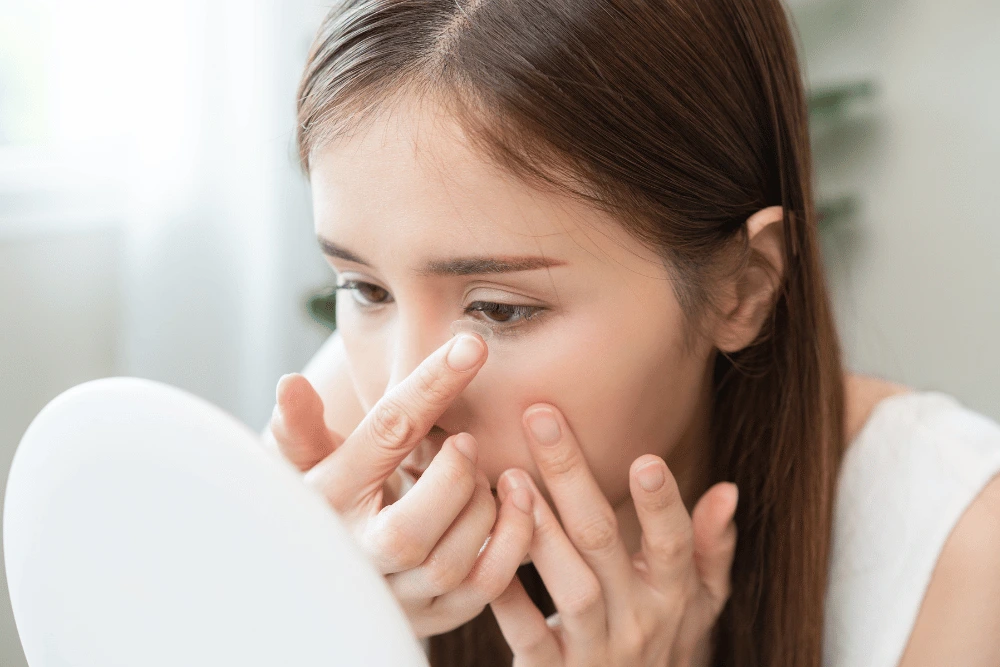 a young woman putting in a contact lens while looking in a mirror