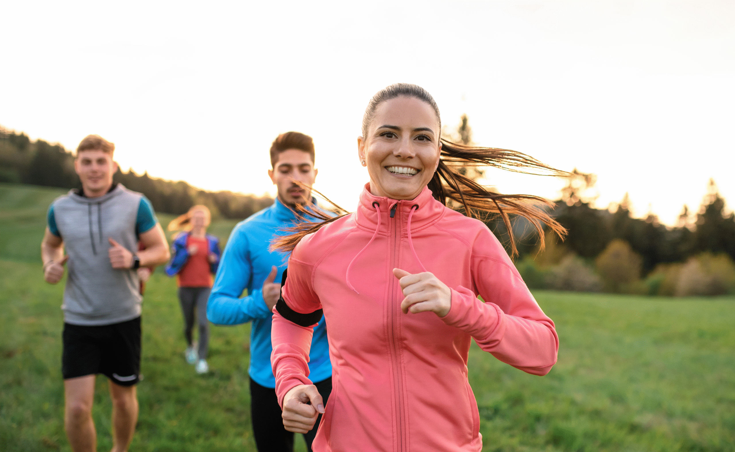 A smiling woman in a pink windbreaker jogs through a green field. Two men and a second woman are jogging in the background behind her.